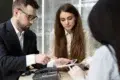 Three professionals in a meeting review documents on a tablet and phone.