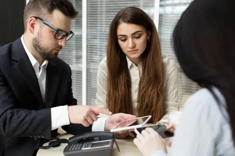 Three professionals in a meeting review documents on a tablet and phone.