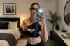 A young woman in a black cutout top and sunglasses takes a mirror selfie in a bedroom with a framed city skyline print.