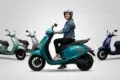 Woman in a helmet riding a teal Chetak electric scooter, with other scooters in various colors behind her.