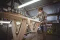 Craftsman wearing a mask sands a wooden surfboard blank in a workshop.