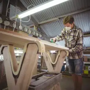 Craftsman wearing a mask sands a wooden surfboard blank in a workshop.
