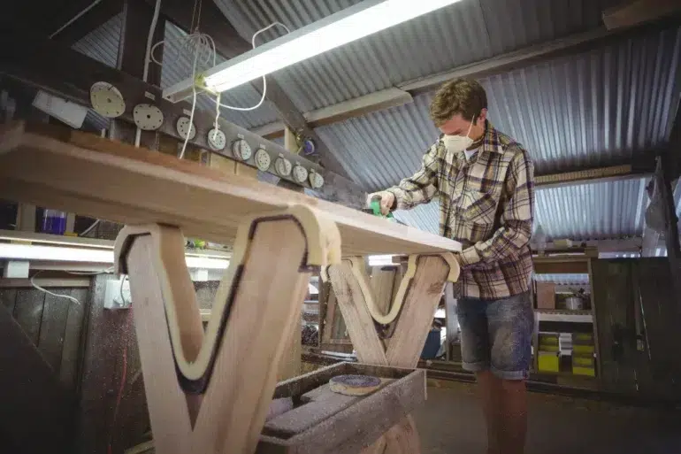 Craftsman wearing a mask sands a wooden surfboard blank in a workshop.