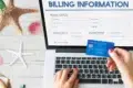 Hands hold a blue credit card over a laptop displaying an online billing form. A starfish and seashells are on the white wooden surface, suggesting a beach theme.
