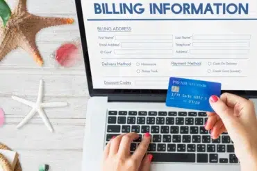 Hands hold a blue credit card over a laptop displaying an online billing form. A starfish and seashells are on the white wooden surface, suggesting a beach theme.