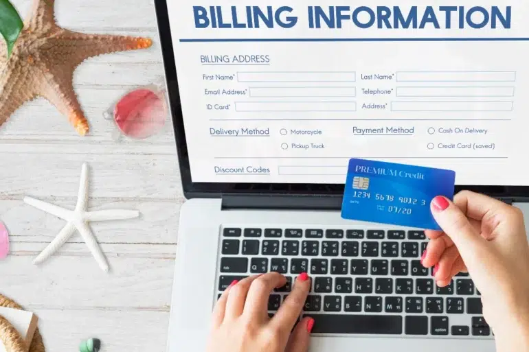Hands hold a blue credit card over a laptop displaying an online billing form. A starfish and seashells are on the white wooden surface, suggesting a beach theme.