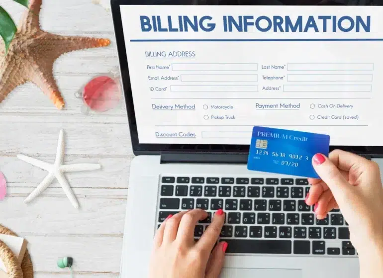 Hands hold a blue credit card over a laptop displaying an online billing form. A starfish and seashells are on the white wooden surface, suggesting a beach theme.