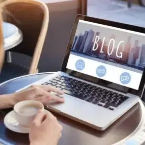 Woman working on a laptop with the word "BLOG" displayed on screen, a cup of coffee beside her.