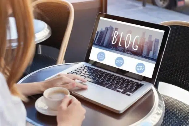 Woman working on a laptop with the word "BLOG" displayed on screen, a cup of coffee beside her.