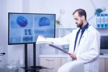 Doctor in lab coat reviews medical chart in front of screen displaying brain scans and DNA helix.