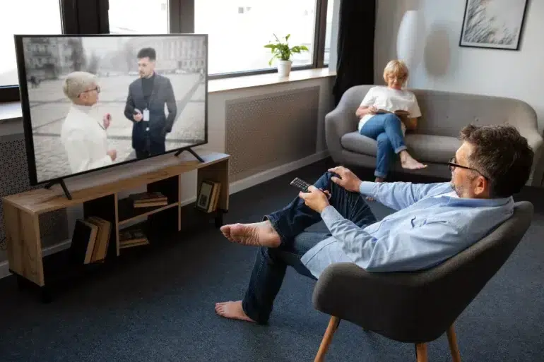 A man in glasses sits in a modern living room watching TV, holding a remote. A woman relaxes on a sofa, reading. The atmosphere is calm and cozy.