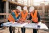 Three construction workers in hard hats and safety vests review blueprints at a construction site.