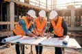 Three construction workers in hard hats and safety vests review blueprints at a construction site.