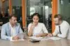 Three colleagues in a meeting room, engaged in discussion. One man holds a tablet, while the woman and another man look thoughtful. The mood is focused.