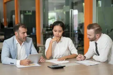 Three colleagues in a meeting room, engaged in discussion. One man holds a tablet, while the woman and another man look thoughtful. The mood is focused.