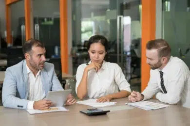 Three colleagues in a meeting room, engaged in discussion. One man holds a tablet, while the woman and another man look thoughtful. The mood is focused.