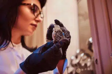 A person wearing black gloves examines a luxury watch under warm lighting. The focus is on the fine details of the watch, suggesting elegance and precision.