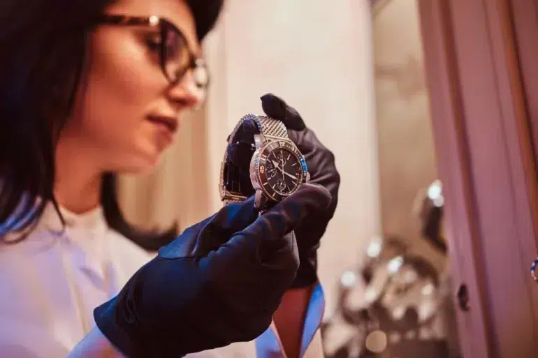A person wearing black gloves examines a luxury watch under warm lighting. The focus is on the fine details of the watch, suggesting elegance and precision.