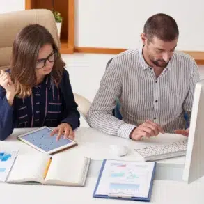 A man and woman work together at a desk. The woman reviews charts on a tablet, while the man types on a keyboard. Documents and a coffee cup are on the desk.