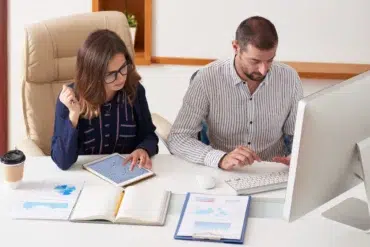 A man and woman work together at a desk. The woman reviews charts on a tablet, while the man types on a keyboard. Documents and a coffee cup are on the desk.