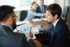 Two businessmen in suits discuss a bar graph on a clipboard in a meeting.