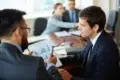 Two businessmen in suits discuss a bar graph on a clipboard in a meeting.