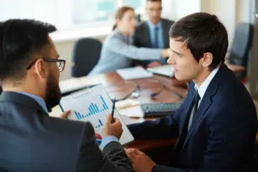 Two businessmen in suits discuss a bar graph on a clipboard in a meeting.
