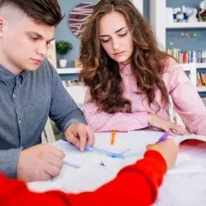 Three students study together at a table with books and pens.