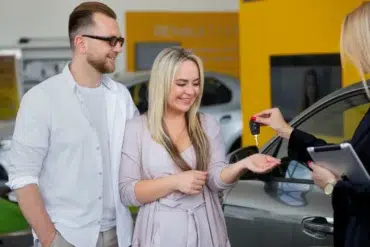 A smiling couple receives car keys from a salesperson in a dealership. The atmosphere is joyful and celebratory, highlighting a successful purchase.