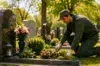 A gardener in green overalls kneels beside a grave, tending to pink and white flowers. Sunlight filters through trees, creating a serene cemetery scene.