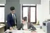 Two business professionals in an office, one standing holding papers, the other sitting and writing at a desk with documents and a printer nearby.