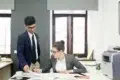 Two business professionals in an office, one standing holding papers, the other sitting and writing at a desk with documents and a printer nearby.