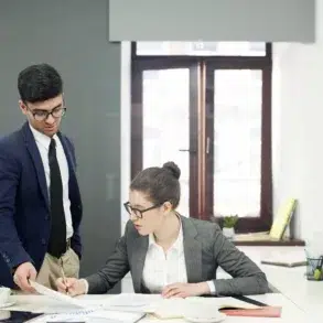 Two business professionals in an office, one standing holding papers, the other sitting and writing at a desk with documents and a printer nearby.