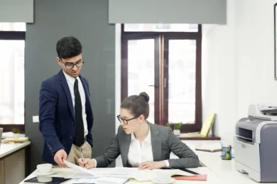 Two business professionals in an office, one standing holding papers, the other sitting and writing at a desk with documents and a printer nearby.
