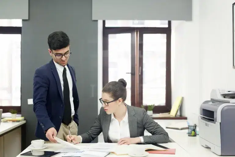 Two business professionals in an office, one standing holding papers, the other sitting and writing at a desk with documents and a printer nearby.