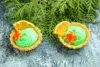 Two tarts with green cream, orange slices, and star-shaped cookies sit on a textured blue surface in front of green evergreen branches.