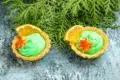 Two tarts with green cream, orange slices, and star-shaped cookies sit on a textured blue surface in front of green evergreen branches.