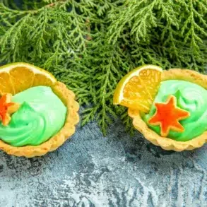 Two tarts with green cream, orange slices, and star-shaped cookies sit on a textured blue surface in front of green evergreen branches.