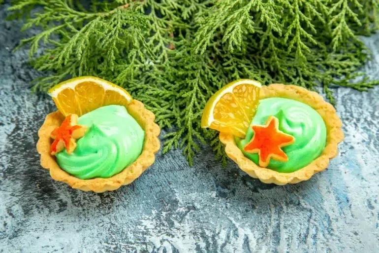 Two tarts with green cream, orange slices, and star-shaped cookies sit on a textured blue surface in front of green evergreen branches.