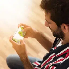 A man in a striped shirt intently plays a game on his phone. The wooden floor and casual setting create a relaxed atmosphere.