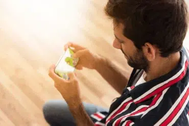 A man in a striped shirt intently plays a game on his phone. The wooden floor and casual setting create a relaxed atmosphere.
