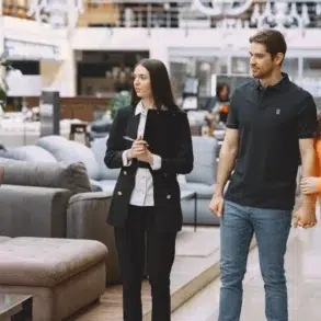 A couple holds hands while looking at sofas in a furniture store. A salesperson with a clipboard guides them, showcasing a professional and welcoming atmosphere.