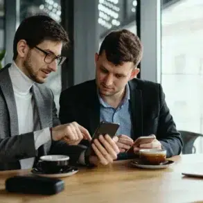 Two men sit at a wooden table in a cafe, engaged in conversation. One points at a smartphone while the other listens, both holding coffee cups.