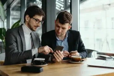 Two men sit at a wooden table in a cafe, engaged in conversation. One points at a smartphone while the other listens, both holding coffee cups.