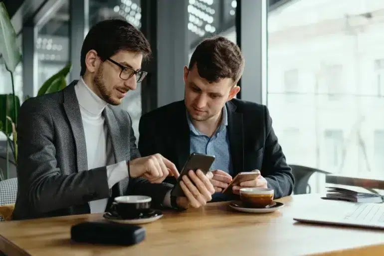 Two men sit at a wooden table in a cafe, engaged in conversation. One points at a smartphone while the other listens, both holding coffee cups.