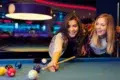 Two women playing pool in a dimly lit billiards room, one aiming to strike the cue ball with a pool stick.