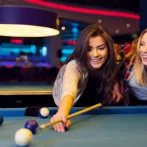 Two women playing pool in a dimly lit billiards room, one aiming to strike the cue ball with a pool stick.