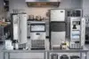 Four stainless steel ice makers on a kitchen counter, filled with ice cubes. The setting is industrial, with a stove and cookware in the background.