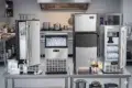 Four stainless steel ice makers on a kitchen counter, filled with ice cubes. The setting is industrial, with a stove and cookware in the background.