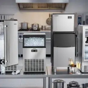 Four stainless steel ice makers on a kitchen counter, filled with ice cubes. The setting is industrial, with a stove and cookware in the background.